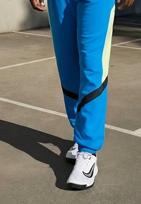 Persona che indossa pantaloni della tuta blu brillante e scarpe da ginnastica bianche Nike mentre cammina su un campo all'aperto segnato.