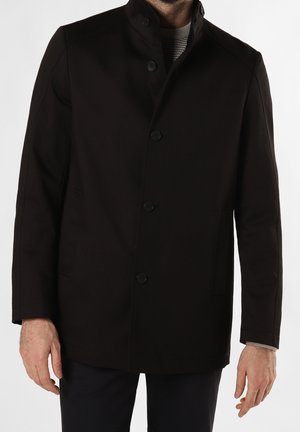 Hombre vestido con un abrigo negro abotonado con cuello alto y bolsillos laterales, combinado con pantalones oscuros y un suéter gris debajo.