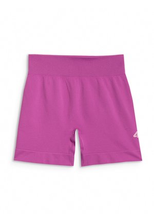 Młoda dziewczyna stoi, ubrana w fioletowy sportowy crop top i szorty z białymi skarpetkami i fioletowymi trampkami na szarym tle.