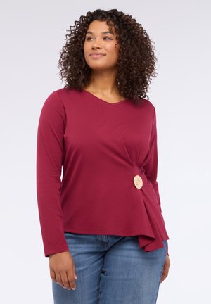 Maglia lunga bordeaux con scollo a V e design asimmetrico, caratterizzata da un elegante bottone dorato decorativo sul lato. Indossata con jeans blu denim.