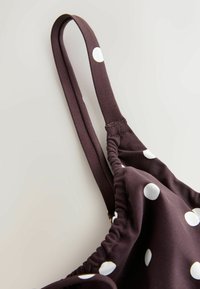 Pruun kangas valgete täppidega, millel on õhukesed riputusrihmad ja volditud detailid. Sile tekstuur, osaline vaade küljelt.