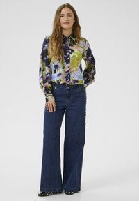 Večbarvna srajca s kratkimi in dolgimi rokavi v tehniki tie-dye, v kombinaciji s temno modrimi širokimi hlačami z dodatki s kovinskimi studii ob straneh.