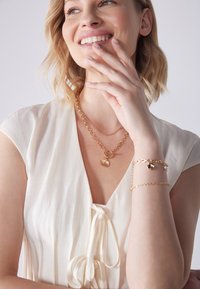 Gelaagde gouden ketting met een schelp hanger, gecombineerd met een delicate gouden kettingarmband. Model draagt een lichtcrème blouse met een strikdetail.