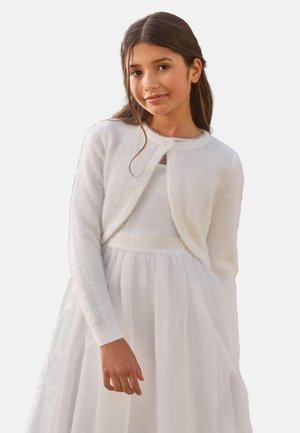 Dziewczyna z długimi brązowymi włosami, ubrana w biały puchaty sweter z guziczkami na białej sukience bez rękawów, stojąca na jednolitym tle.