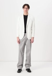 Weißer Blazer über einem schwarzen T-Shirt, grauen Hose und schwarzen Schuhen. Der Blazer hat Taschen und eine taillierte Passform.