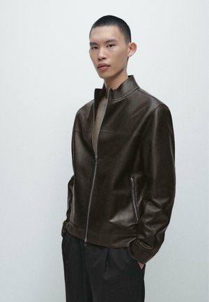 Młody mężczyzna o krótkich włosach, ubrany w ciemnobrązową skórzaną kurtkę zapinaną na suwak i czarne spodnie, stojący na tle jednolitej jasnej ściany.