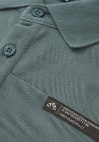 Koszulka polo w stonowanym zielonym kolorze, z teksturowanym wzorem, dwoma ciemnymi guzikami oraz czarną metką z białym tekstem i logo.