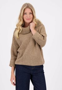 Beige chunky gebreide trui met een grote geribbelde kraag, driekwartmouwen en een relaxte pasvorm, gecombineerd met donkerblauwe jeans.