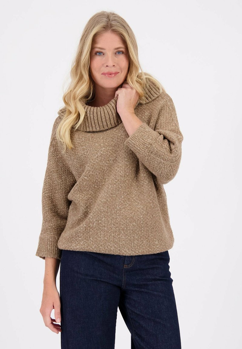 Beige chunky gebreide trui met een grote geribbelde kraag, driekwartmouwen en een relaxte pasvorm, gecombineerd met donkerblauwe jeans.