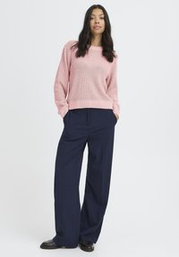 Femme debout avec les mains dans les poches, portant un pull en tricot rose, un pantalon large bleu marine et des chaussures noires sur un fond blanc.
