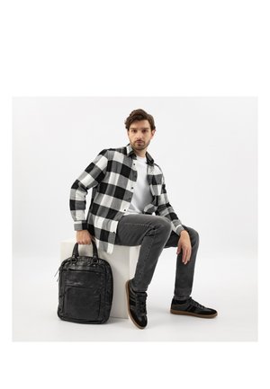 Mężczyzna w czarno-białej koszuli w kratę i szarych spodniach siedzi na białym bloku, trzymając czarny skórzany plecak na białym tle.