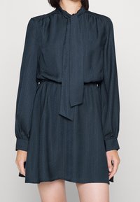 Tummanvihreä, kuviollinen mekko, jossa pitkät hihat, korkea kaulus ja solmimisdetaili. Pehmeä, teksturoitu kangas ja joustava vyötärö rennon istuvuuden takaamiseksi.