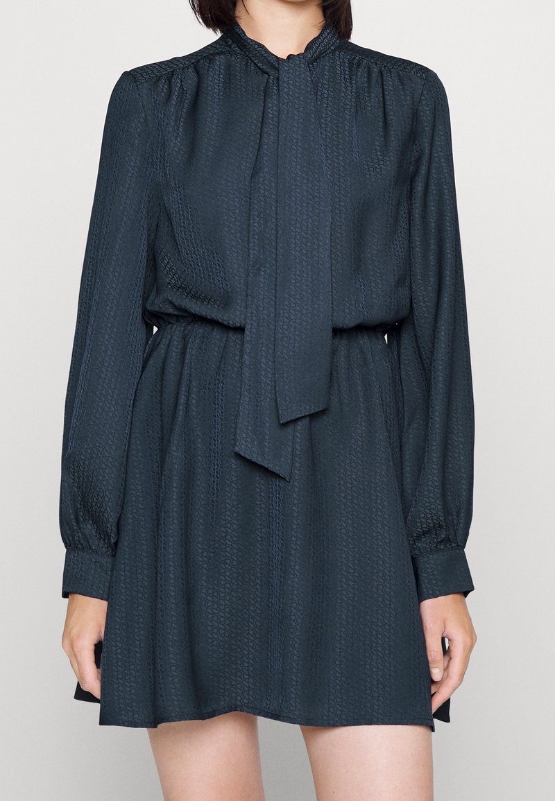 Tummanvihreä, kuviollinen mekko, jossa pitkät hihat, korkea kaulus ja solmimisdetaili. Pehmeä, teksturoitu kangas ja joustava vyötärö rennon istuvuuden takaamiseksi.