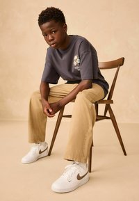 Tricou gri cu un grafic de craniu și trandafir, pantaloni khaki și adidași albi Nike cu accente maronii, așezat pe un scaun din lemn, pe un fundal bej.