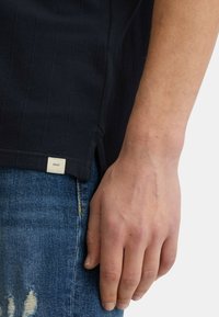 Polo shirt navy con righe verticali, maniche corte e spacco laterale; etichetta vicino all'orlo. Abbinato a jeans blu sbiaditi.