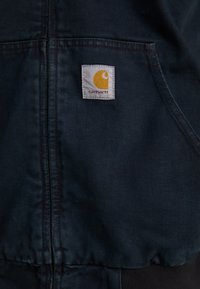 Dark blue Carhartt jacket with front zipper and kangaroo pocket, featuring a small white patch with yellow logo and red text on chest.