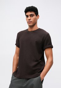 Brązowa koszulka z krótkim rękawem, okrągłym dekoltem i podwiniętymi mankietami, wykonana z teksturowanej tkaniny, w zestawieniu z szarymi spodniami.