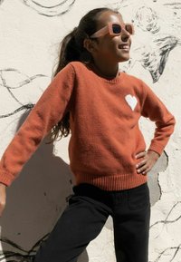 Pull en couleur rouille avec un ourlet et des poignets côtelés; présente un motif de cœur blanc. Porté avec un pantalon noir et des lunettes de soleil transparentes contre un mur.