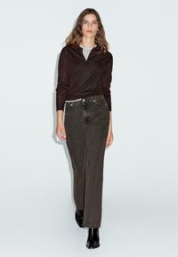 Dunkelbraunes Langarmoberteil, das über einem grauen Hemd getragen wird, kombiniert mit hoch sitzenden, olivgrünen Straight-Leg-Jeans und schwarzen Stiefeletten.