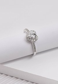 Zilveren ring met een hartvormige centrale steen omringd door kleinere stenen. De band heeft een gladde afwerking en délicate details.