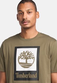 Olivově zelené bavlněné tričko s centrálním bílým čtvercovým grafickým návrhem se vzorem stromu a nápisem "Timberland" tučným černým písmem.