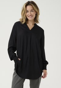 Túnica negra con un patrón de diamantes, escote en V, mangas largas con puños fruncidos y bolsillos laterales, combinada con pantalones grises.