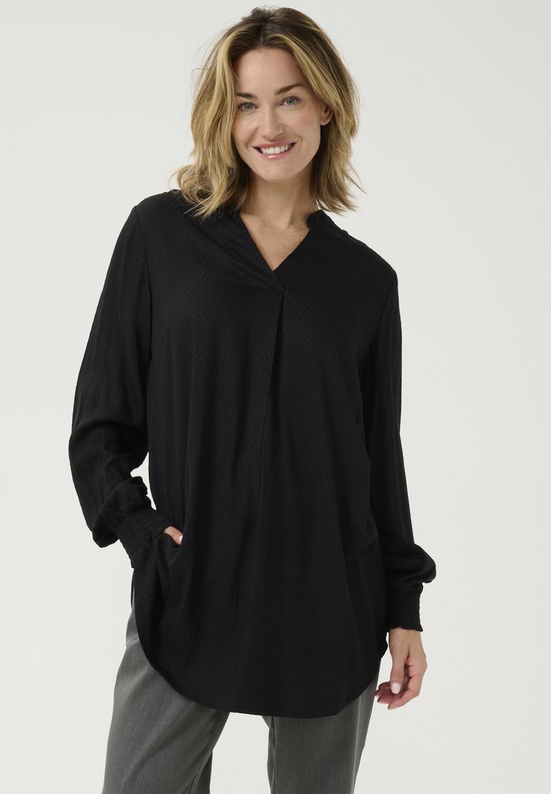 Túnica negra con un patrón de diamantes, escote en V, mangas largas con puños fruncidos y bolsillos laterales, combinada con pantalones grises.