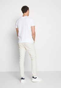 Biała koszulka z krótkim rękawem i dekoltem w kształcie litery V, w połączeniu z jasnobeżowymi spodniami. Buty są białe z granatowymi akcentami. Jednolity tle.