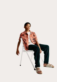 Camicia a maniche corte di colore corallo con motivi floreali neri, maglietta bianca sotto, pantaloni scuri e sandali beige. Seduto su una sedia pieghevole bianca.