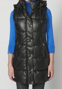 Femme portant un gilet matelassé en cuir noir sans manches avec une capuche, sur un haut bleu à manches longues, mains détendues le long du corps.