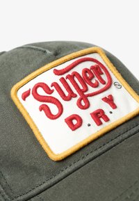 Gorra de algodón verde con un parche bordado blanco que lleva el texto rojo "Super" y "D.R.Y.", rodeado de un borde de costura amarilla, mostrando un diseño estructurado.