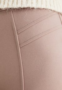 Beige tkaninske hlače z gladko teksturo, ki imajo okrasne šive in prilegan kroj. Del pletenega puloverja je viden zgoraj.