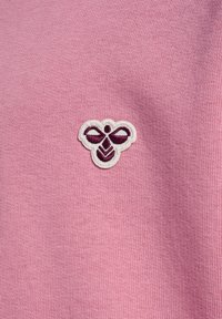 Lille broderet mørkelilla og hvidt bi-logo centreret på blød lyserød strikket stof baggrund.