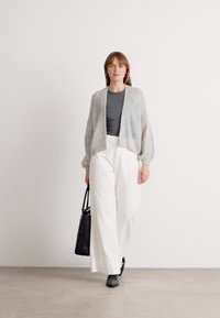 Lichtgrijze gebreide cardigan, grijze T-shirt, witte wijde broek, zwarte schoenen en een zwarte handtas. Model loopt op een effen achtergrond.
