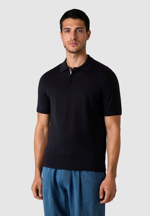 Mand iført en tætsiddende sort poloshirt med korte ærmer og løse blå bukser, stående mod en ensfarvet grå baggrund.