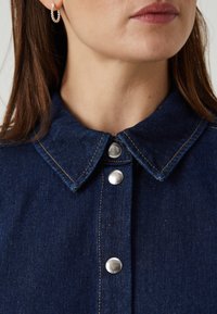 Mörk denimskjorta med en strukturerad krage, silverfärgade tryckknappar och kontrasterande sömmar. Slät textur med en klassisk design.