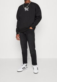Persona con sudadera negra con el logotipo blanco "Bsl", pantalones negros, zapatillas Adidas blancas con rayas negras y una cadena de plata.