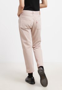 Pantalones tapered rosa claro de algodón con un acabado ligeramente texturizado, que cuentan con bolsillos traseros y costuras distintivas de la marca. Zapatos negros visibles.