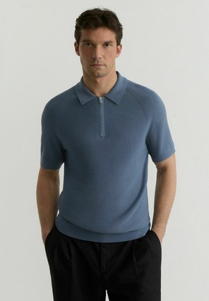 Mees seisab käed taskutes, kandes sinist lühikese varrukaga kampsunist polosärki, millel on ees lukk, ja musti pükse ühevärvilise tausta ees.