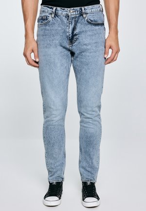 Mand iført lyseblå slim-fit jeans og sorte sneakers, der står mod en ensfarvet lys baggrund.
