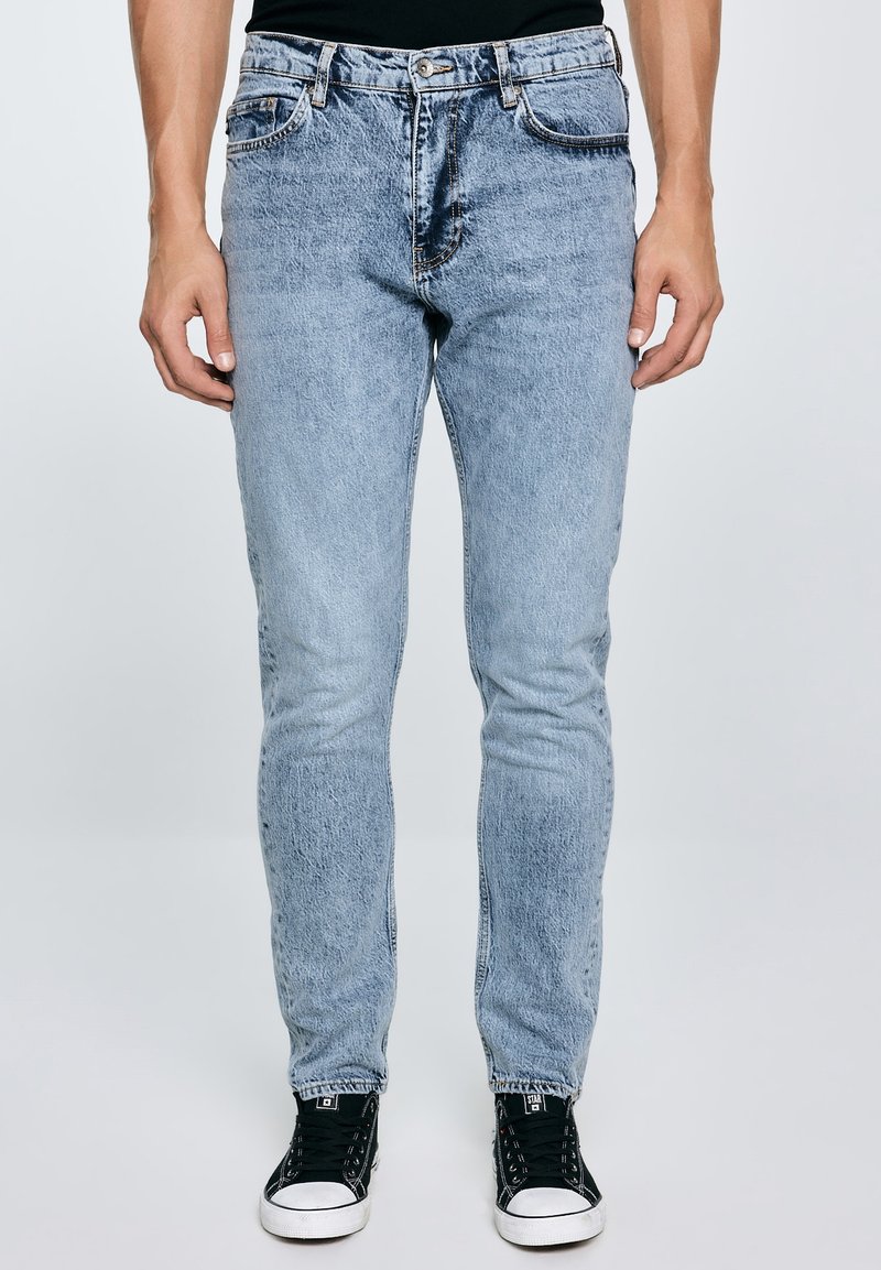 Homme portant un jean slim bleu clair et des baskets noires, debout devant un fond clair uni.