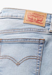 Ljust blå denimjeans med en tanfärgad läderlapp som har röd branding. Jeanssen har orange sömmar och en ok-design ovanför bakfickorna.