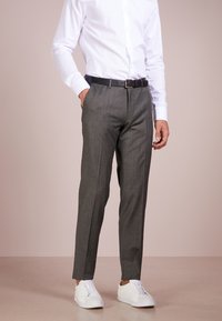 Uomo che indossa pantaloni grigi, camicia bianca, cintura nera e sneaker bianche, in piedi contro uno sfondo chiaro e neutro.