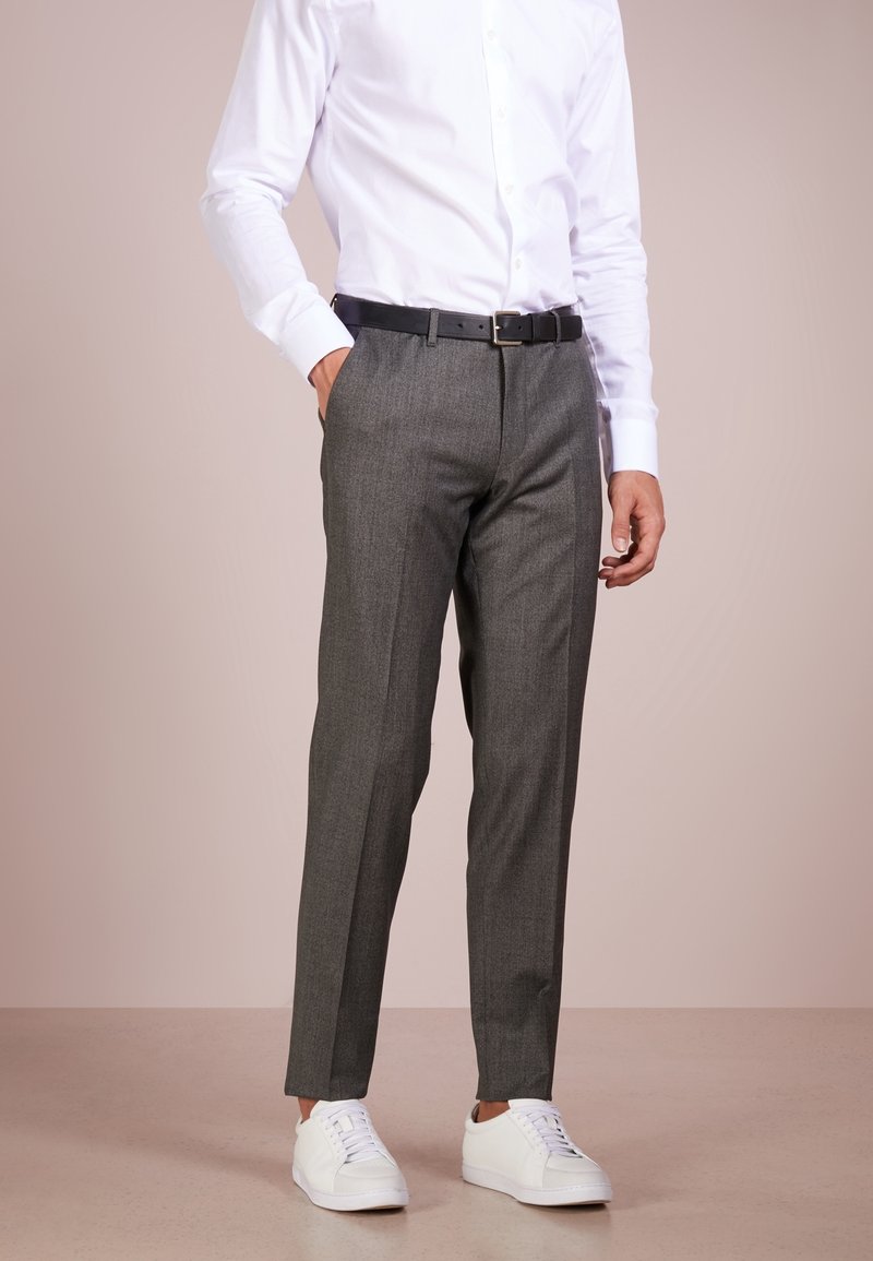 Uomo che indossa pantaloni grigi, camicia bianca, cintura nera e sneaker bianche, in piedi contro uno sfondo chiaro e neutro.