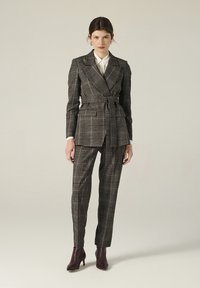 Donna che indossa un blazer a quadri marrone con cintura, pantaloni abbinati e stivali alla caviglia marroni scuri, in piedi di fronte a uno sfondo semplice.