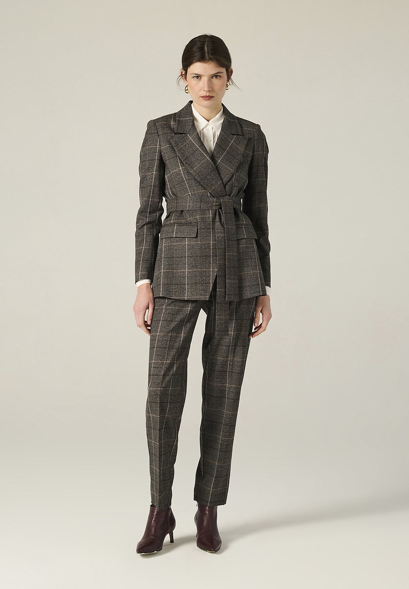 Donna che indossa un blazer a quadri marrone con cintura, pantaloni abbinati e stivali alla caviglia marroni scuri, in piedi di fronte a uno sfondo semplice.