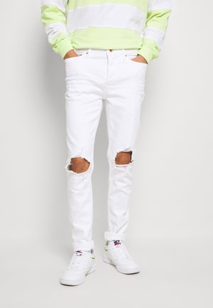 Personne portant un jean blanc déchiré avec des trous aux genoux, des baskets blanches et un haut à manches longues rayé vert clair et blanc.