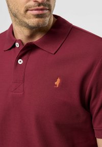 Bordo polo marškiniai su tekstūruota medžiaga, trimis mygtukais priekyje ir išsiuvinėtu oranžiniu logotipu kairėje krūtinės pusėje.