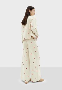 Beige denim outfit met een losvallende jas en wijdvallende broek, beide versierd met kleine rode bloemenpatronen en het woord "SOL."