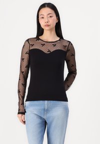 Top negro de manga larga con un cuerpo ajustado y mangas de encaje transparente con puntos que presentan patrones de lazos. Escote suave y tejido suave.